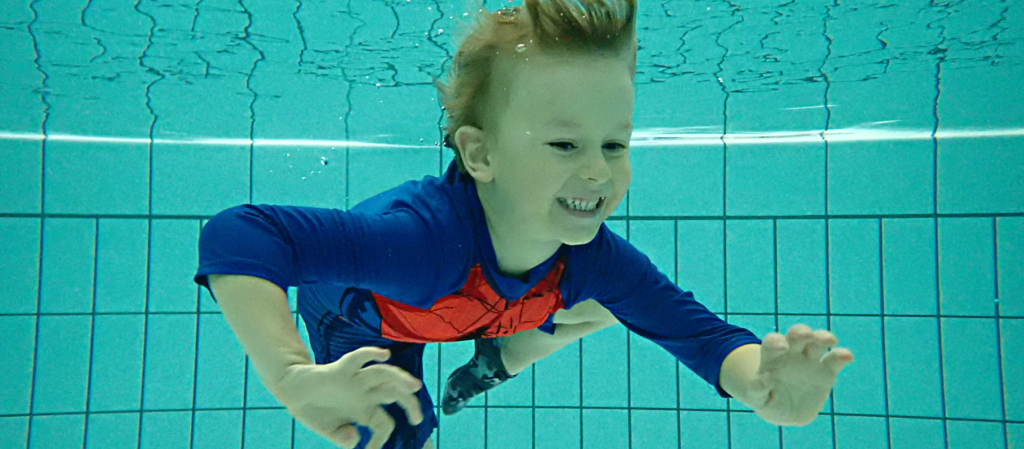 Pieni poika sukeltaa hymyillen altaassa hämähäkkimies uima-asussa.
