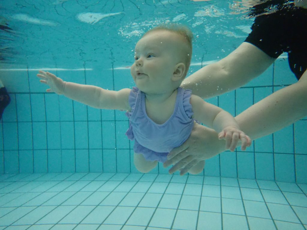 Vauva vaaleanvioletissa uimapuvussa sukeltaa veden alla altaassa aikuisen pitäessä kevyesti kiinni. 